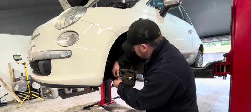 Mechanic unplugging the brake pad wear sensor on a Fiat 500 with the front wheel removed, exposing the disc and caliper